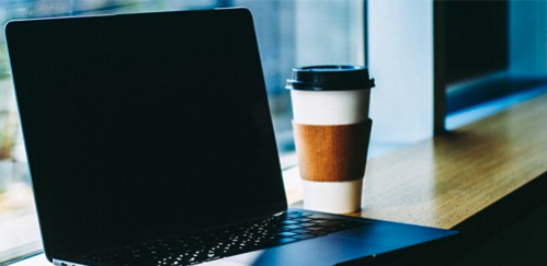 A laptop with a dark screen sits on a wooden table next to a disposable coffee cup with a cardboard sleeve, by a window with natural light shining in.