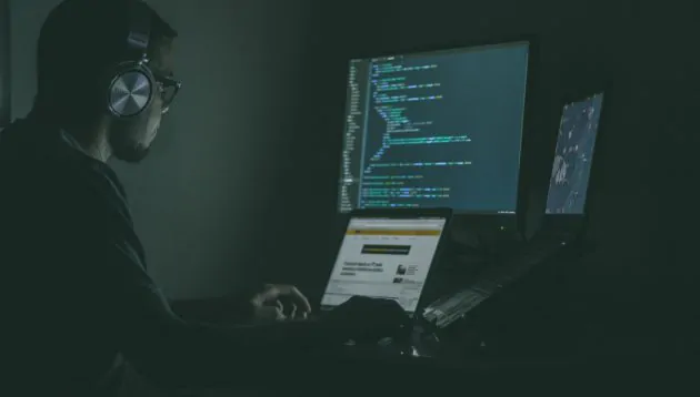 A person wearing headphones sits in a dimly lit room, working on a laptop with code—such as redux middleware—displayed on two monitors. The main screen shows programming code, whilst another screen displays a webpage.